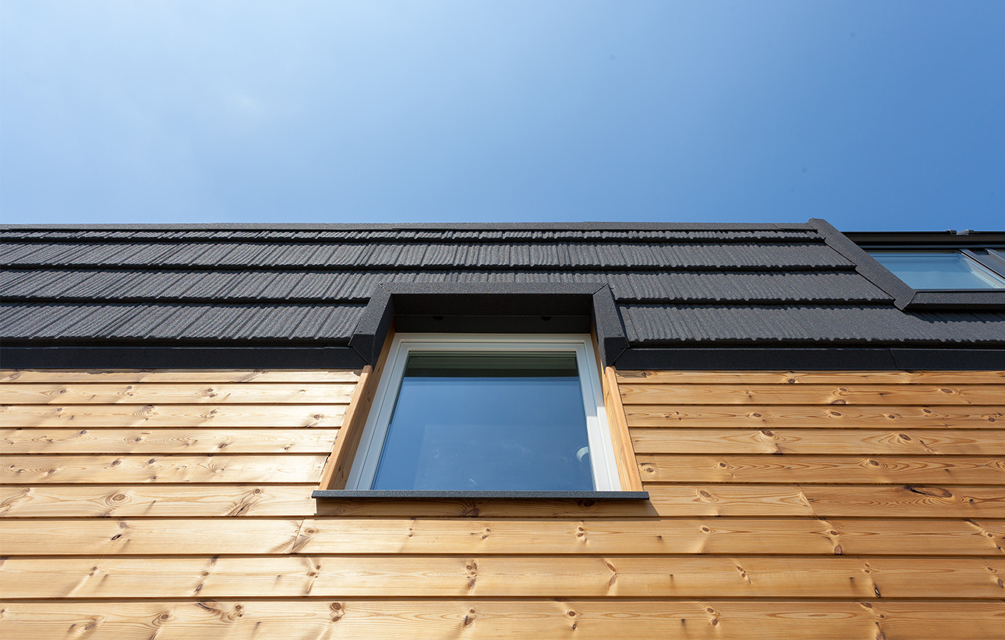 Nahaufnahme einer modernen Holzfassade mit Fenster und schwarzem Dachabschluss bei strahlend blauem Himmel.