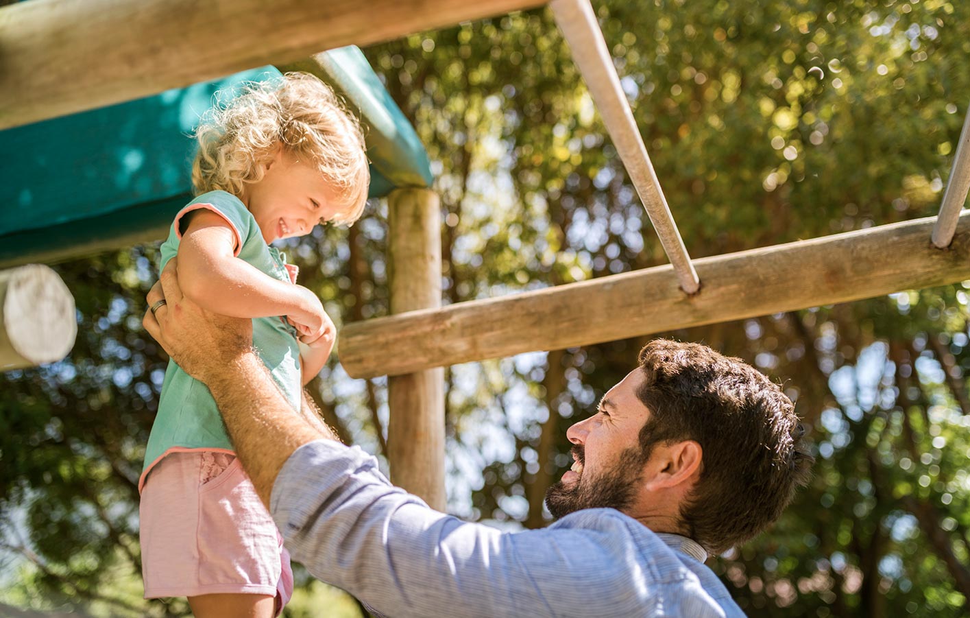 Vater hebt kleines Kind an einem Spielgerät aus Holz im Garten hoch